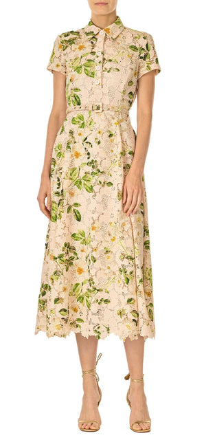 A woman wears the Monique Lhuillier Lace Polo Shirt Dress in beige with rose print, guipure lace, short sleeves, collar, matching belt, and a tea-length A-line skirt, paired with strappy beige heels.