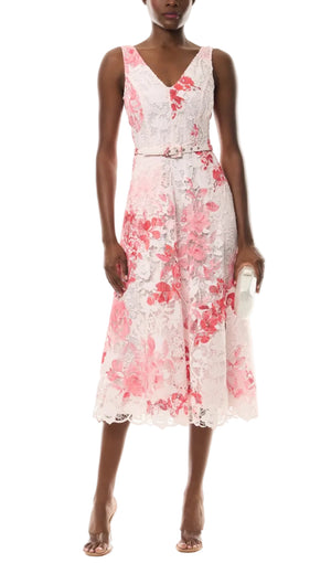 A woman wears the Monique Lhuillier Rose Printed Lace Midi Dress, featuring white lace with pink florals, styled with a small clutch and open-toe heels.
