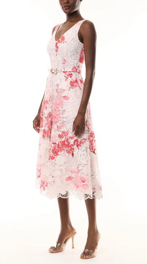 A woman wears the Monique Lhuillier Rose Printed Lace Midi Dress, belted at the waist for a feminine silhouette, paired with nude open-toe heels.