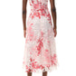 A person stands facing away, wearing the Monique Lhuillier Rose Printed Lace Midi Dress in white with pink and red floral patterns, styled with nude heeled sandals.