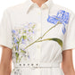 A woman wears the Monique Lhuillier Iris Printed Poplin Day Dress in Silk White, featuring a collared neckline, button front, matching belt, and blue and green floral prints on the chest.