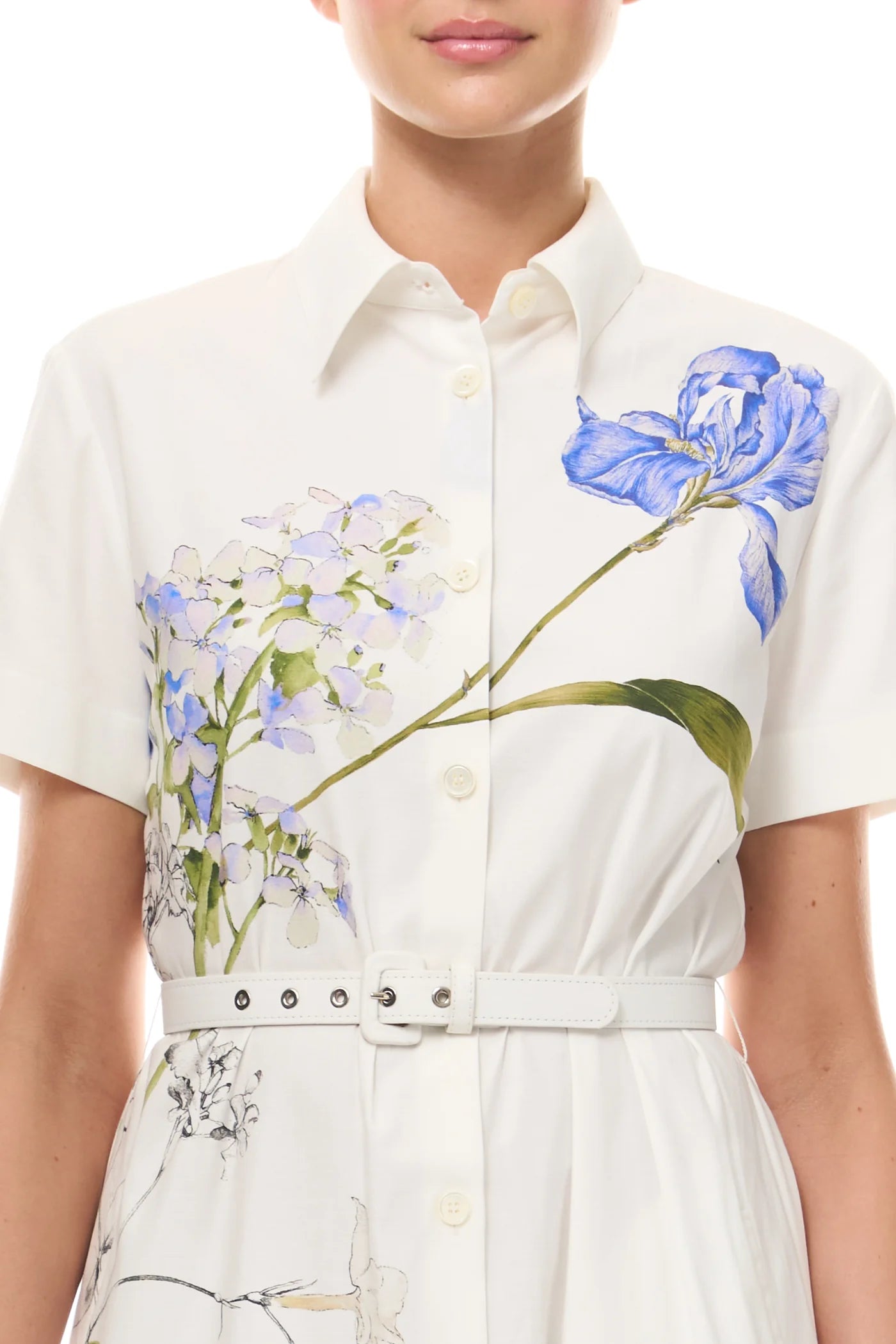 A woman wears the Monique Lhuillier Iris Printed Poplin Day Dress in Silk White, featuring a collared neckline, button front, matching belt, and blue and green floral prints on the chest.