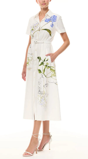 A woman wears the Monique Lhuillier Iris Printed Poplin Day Dress, featuring short sleeves, a button-down front, belted waist, and styled with white high-heeled sandals.