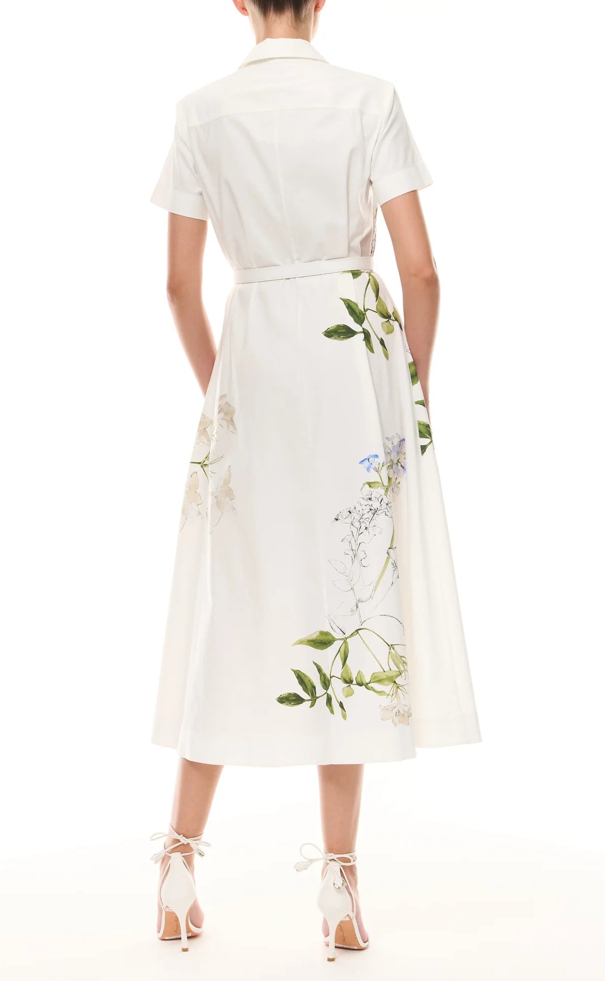 A woman stands facing away, wearing the Monique Lhuillier Iris Printed Poplin Day Dress in Silk White with short sleeves, collar, belted waist, and floral print, paired with white lace-up heels.
