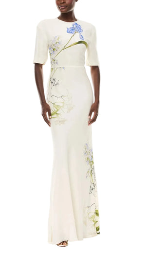 A woman wearing the Iris Bouquet Gown by Monique Lhuillier, a fitted, floor-length dress featuring blue and green floral details on ivory crepe with short sleeves.