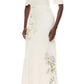 A woman wears the Iris Bouquet Gown by Monique Lhuillier, a short sleeve ivory dress with a long silhouette and blue floral prints, standing against a plain white background.