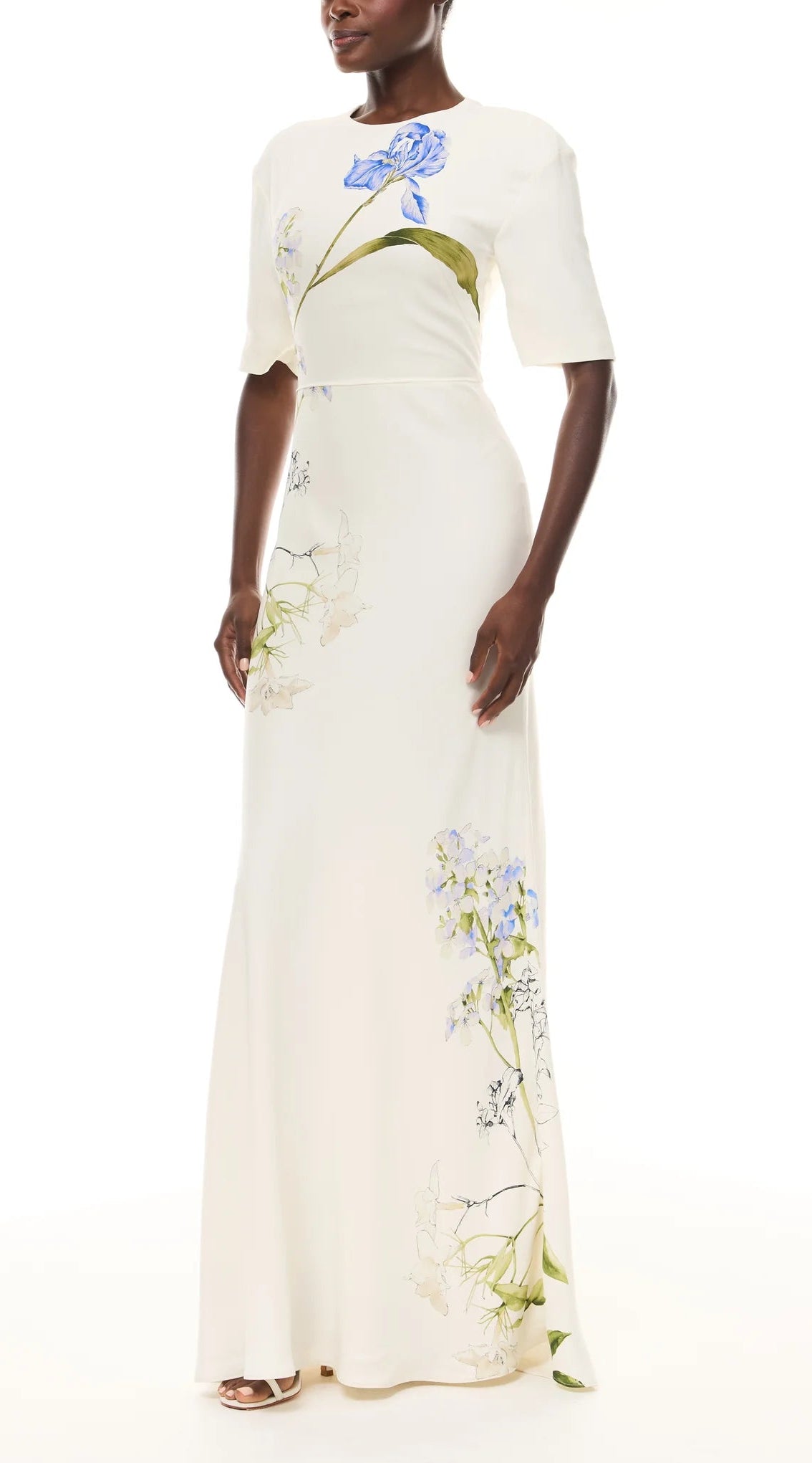 A woman wears the Iris Bouquet Gown by Monique Lhuillier, a short sleeve ivory dress with a long silhouette and blue floral prints, standing against a plain white background.