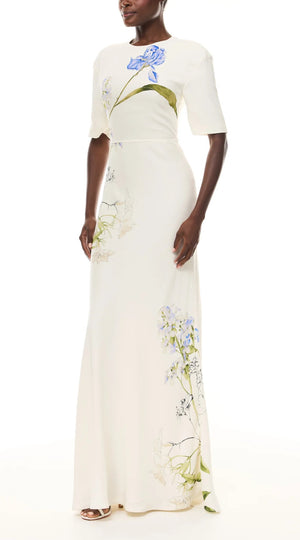 A woman wears the Iris Bouquet Gown by Monique Lhuillier, a short sleeve ivory dress with a long silhouette and blue floral prints, standing against a plain white background.
