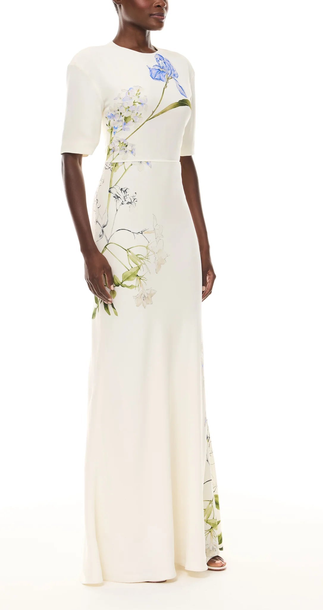 A woman wears the Monique Lhuillier Iris Bouquet Gown, a floor-length dress with short sleeves and a fitted waist, crafted from blue and green floral-printed crepe.