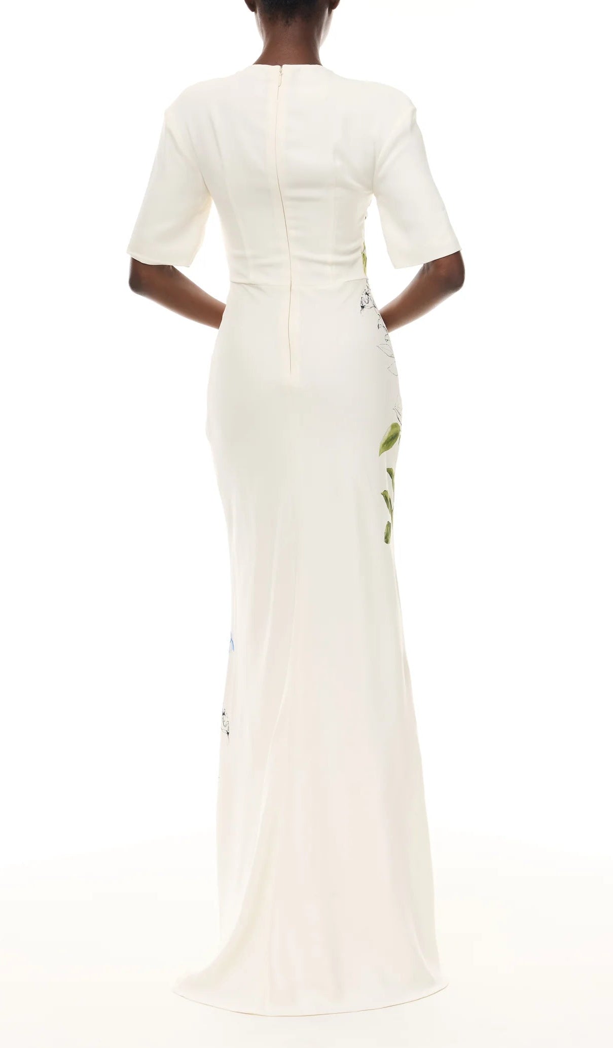 A person wearing the Monique Lhuillier Iris Bouquet Gown stands facing away from the camera against a white background. The fitted, floor-length floral gown features short sleeves and creates an elegant silhouette.