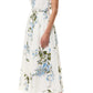 A woman in the Monique Lhuillier Wisteria Floral Poplin Day Dress, featuring a sleeveless design and blue-green floral print, paired with nude high-heeled sandals, stands against a plain background.