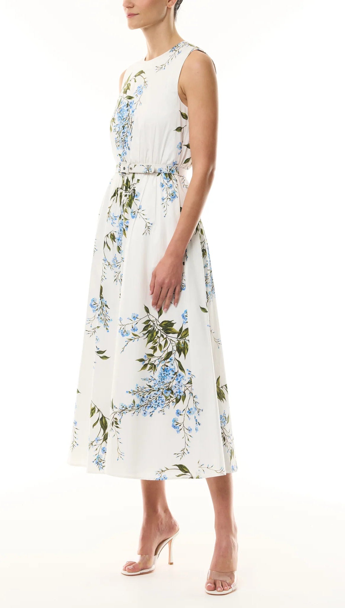 A woman in the Monique Lhuillier Wisteria Floral Poplin Day Dress, featuring a sleeveless design and blue-green floral print, paired with nude high-heeled sandals, stands against a plain background.