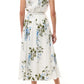 A woman stands with her back to the camera in white heeled sandals, wearing the Monique Lhuillier Wisteria Floral Poplin Day Dress, a sleeveless cotton day dress with blue and green floral print and an A-line skirt.