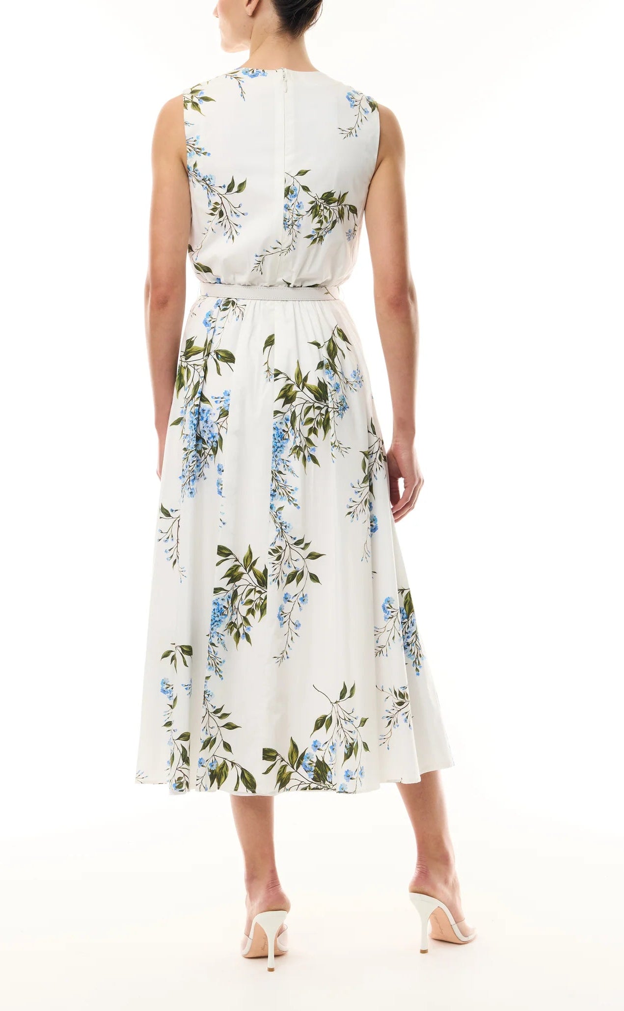 A woman stands with her back to the camera in white heeled sandals, wearing the Monique Lhuillier Wisteria Floral Poplin Day Dress, a sleeveless cotton day dress with blue and green floral print and an A-line skirt.