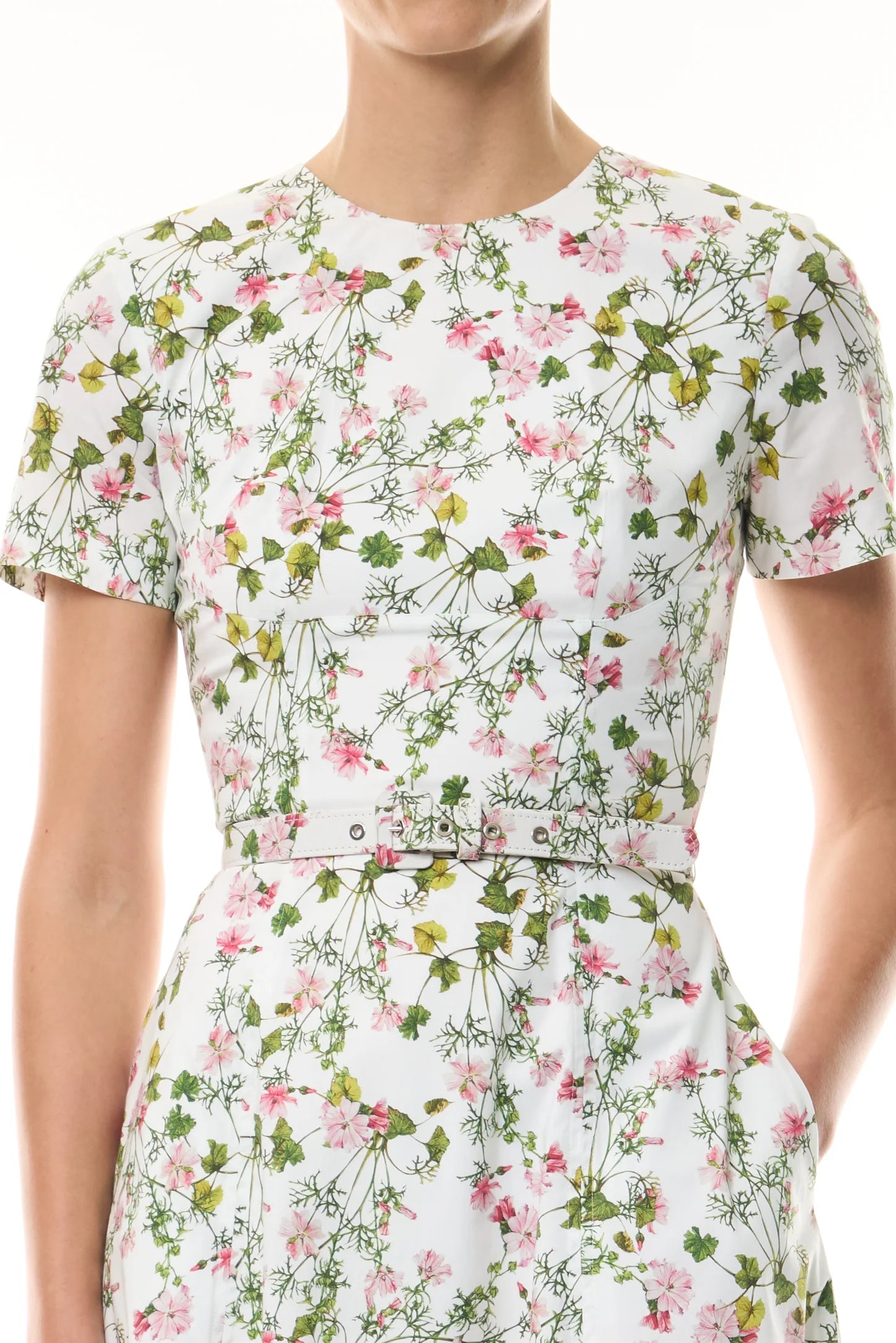 Someone wearing the Monique Lhuillier Ivy Bloom Short Sleeve Floral Day Dress, crafted from white stretch cotton poplin with a pink and green floral print and a coordinating fabric belt.