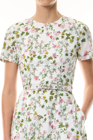 Someone wearing the Monique Lhuillier Ivy Bloom Short Sleeve Floral Day Dress, crafted from white stretch cotton poplin with a pink and green floral print and a coordinating fabric belt.