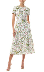 Woman in a Monique Lhuillier Ivy Bloom Short Sleeve Floral Day Dress with pink and green print, nude heels, standing against a white background.