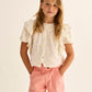 A young girl with long hair stands against a neutral background, wearing the Cozmo Girls' Tansy Floral Blouse—a short-sleeved white cotton gauze top adorned with delicate floral embroidery—and pink pants, her hands in her pockets.