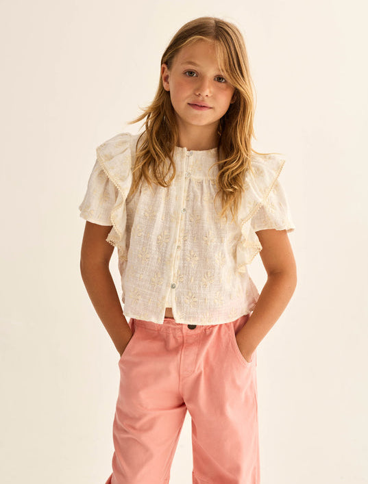 A young girl with long hair stands against a neutral background, wearing the Cozmo Girls' Tansy Floral Blouse—a short-sleeved white cotton gauze top adorned with delicate floral embroidery—and pink pants, her hands in her pockets.