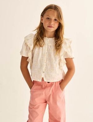 A young girl with long hair stands against a neutral background, wearing the Cozmo Girls' Tansy Floral Blouse—a short-sleeved white cotton gauze top adorned with delicate floral embroidery—and pink pants, her hands in her pockets.