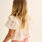 A young girl with long blonde hair, seen from behind, wears the Cozmo Girls' Tansy Floral Blouse and light pink shorts against a plain background.