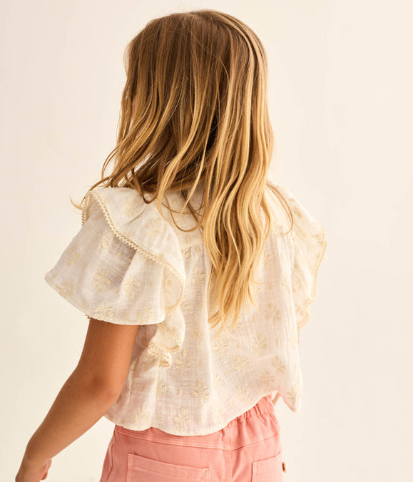 A young girl with long blonde hair, seen from behind, wears the Cozmo Girls' Tansy Floral Blouse and light pink shorts against a plain background.
