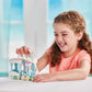 A smiling child in a peach shirt plays with the Calico Critters Floating Cloud Rainbow Train and a small Calico Critters animal figurine while sitting at a table indoors.
