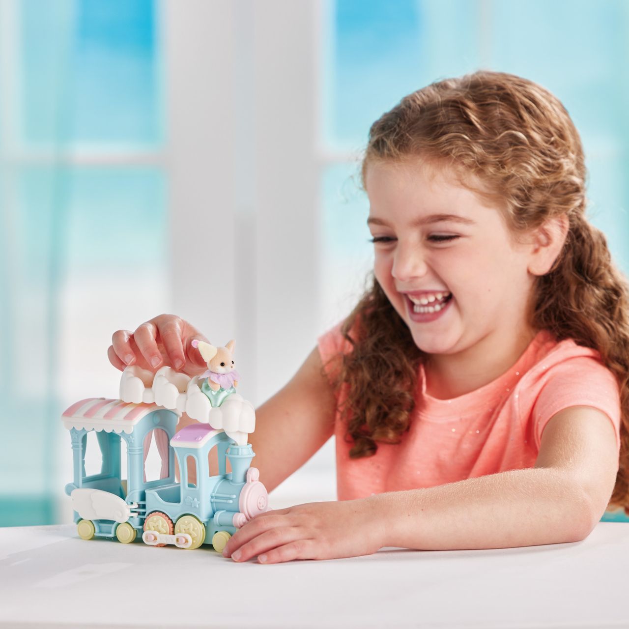 A smiling child in a peach shirt plays with the Calico Critters Floating Cloud Rainbow Train and a small Calico Critters animal figurine while sitting at a table indoors.