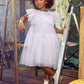 The young girl, capturing a picture-perfect moment, stands with one arm resting on a wooden ladder, dressed in the frilly, light purple Girls’ Ziegfeld Tulle Dress by Tutu du Monde. She pairs it with white socks, sparkly shoes, and a white hair accessory—all set within an indoor rustic setting that evokes timeless Hollywood magic.