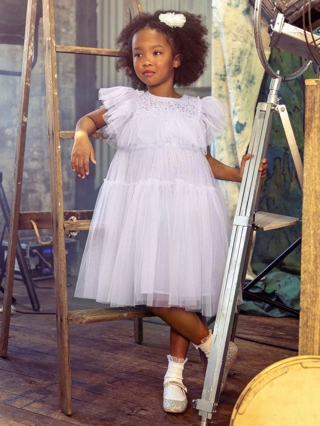 The young girl, capturing a picture-perfect moment, stands with one arm resting on a wooden ladder, dressed in the frilly, light purple Girls’ Ziegfeld Tulle Dress by Tutu du Monde. She pairs it with white socks, sparkly shoes, and a white hair accessory—all set within an indoor rustic setting that evokes timeless Hollywood magic.