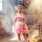 A young girl wears the Tutu du Monde Girls’ Angelina Tutu Dress, featuring ombre pink hues and delicate feathers, as she stands indoors on a wooden floor with vintage film equipment and illuminated marquee lights in the background.