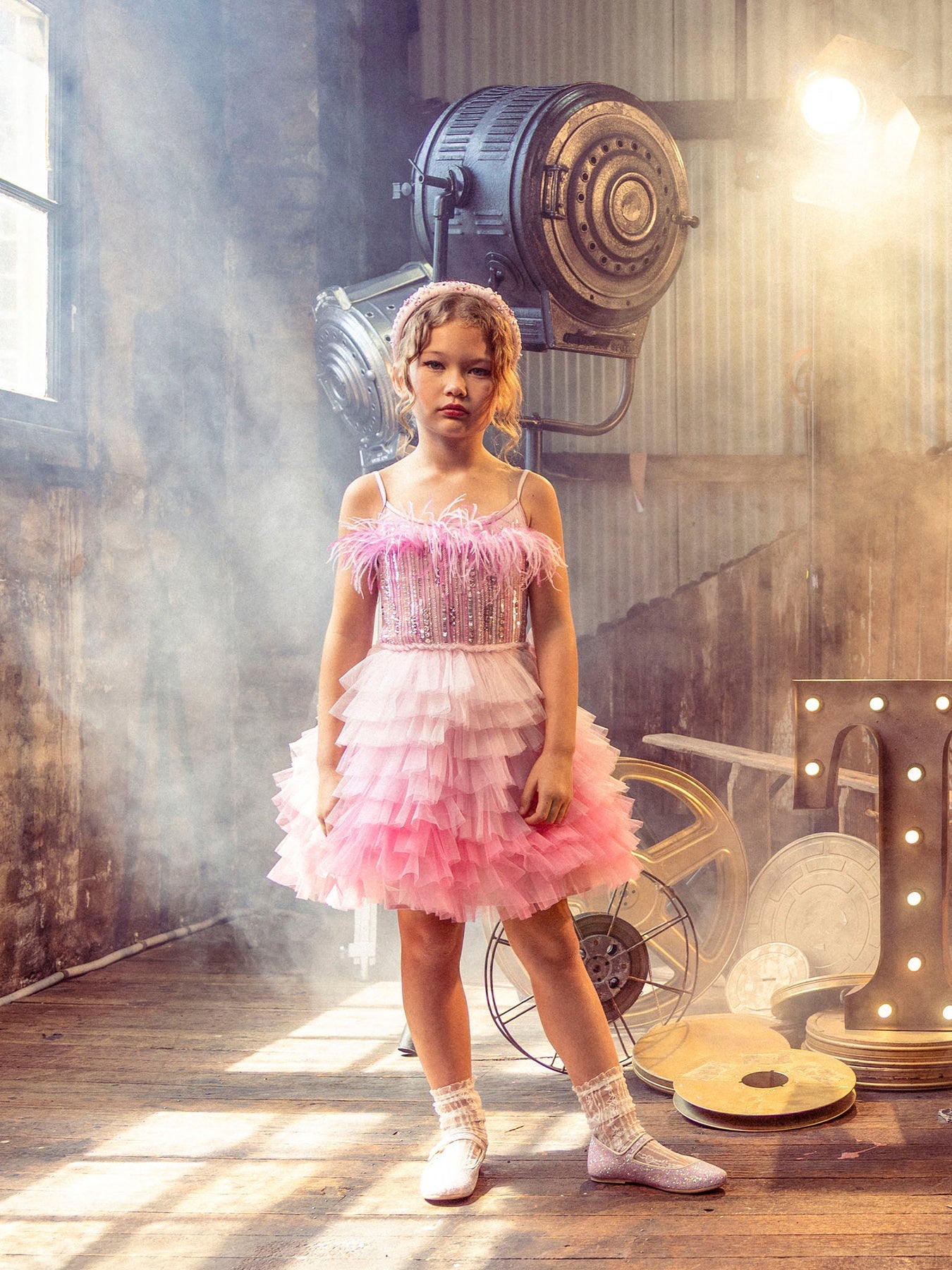 A young girl wears the Tutu du Monde Girls’ Angelina Tutu Dress, featuring ombre pink hues and delicate feathers, as she stands indoors on a wooden floor with vintage film equipment and illuminated marquee lights in the background.