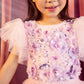 A little girl wearing the Tutu du Monde Girls’ Sparkle in Bloom Tutu Dress in the whimsical Fairy Floss Mix of pinks and purples stands against a pink backdrop, her smile gentle. The enchanting dress features delicate purple and white floral appliqués and shimmering sequins that sparkle beautifully in the light.