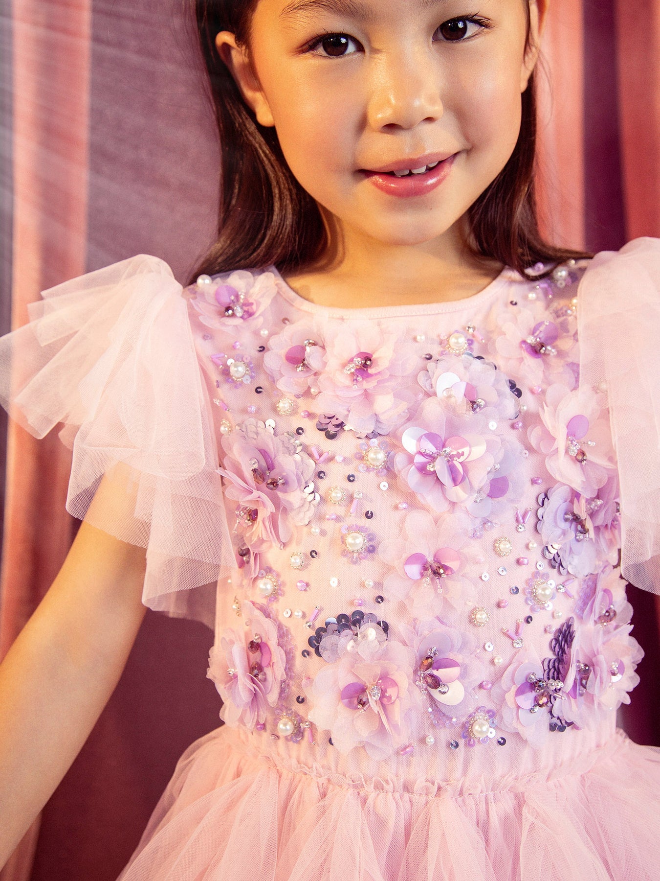 A little girl wearing the Tutu du Monde Girls’ Sparkle in Bloom Tutu Dress in the whimsical Fairy Floss Mix of pinks and purples stands against a pink backdrop, her smile gentle. The enchanting dress features delicate purple and white floral appliqués and shimmering sequins that sparkle beautifully in the light.