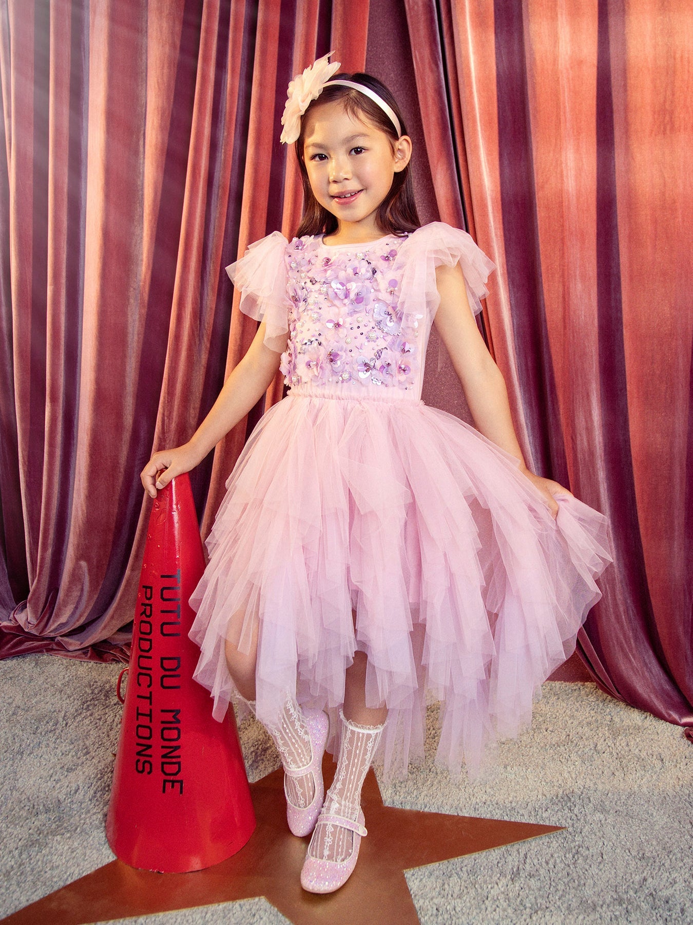 A young girl in a Girls’ Sparkle in Bloom Tutu Dress by Tutu du Monde, with a matching headband, stands on a star-shaped spot holding a red cone labeled “Tutu du Monde Productions” against a curtain backdrop.