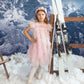 A girl in a Summit Sparkle Tulle Dress by Tutu du Monde stands in faux snow beside crossed skis, framed by snowy mountains.