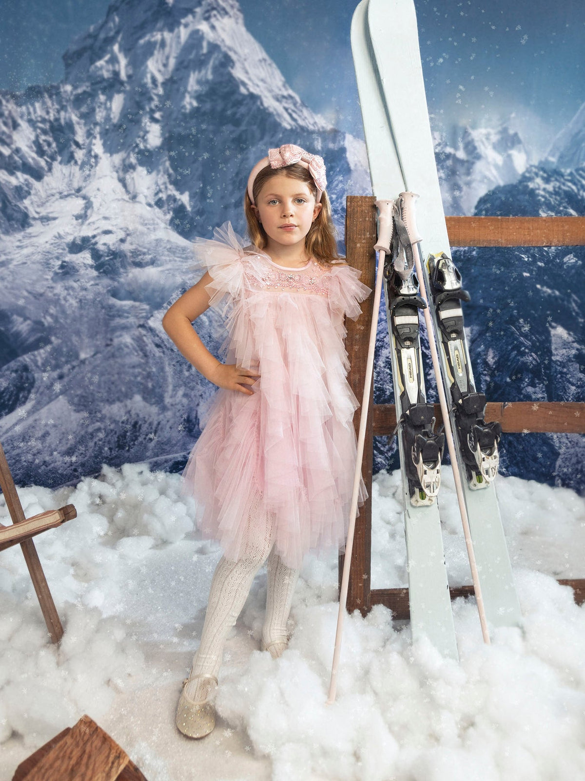 A girl in a Summit Sparkle Tulle Dress by Tutu du Monde stands in faux snow beside crossed skis, framed by snowy mountains.