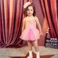 A young child wears the Tutu du Monde Girls’ Bebe Shirley Sparkle Tutu in a peach blush hue, paired with white shoes, standing atop a star-shaped floor mat with a film clapperboard and pink curtains behind—an enchanting scene for little girls who love to shine.