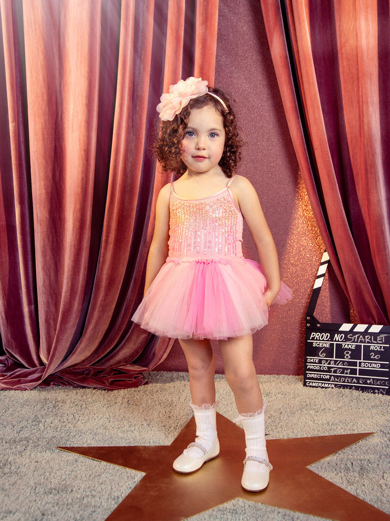 A young child wears the Tutu du Monde Girls’ Bebe Shirley Sparkle Tutu in a peach blush hue, paired with white shoes, standing atop a star-shaped floor mat with a film clapperboard and pink curtains behind—an enchanting scene for little girls who love to shine.