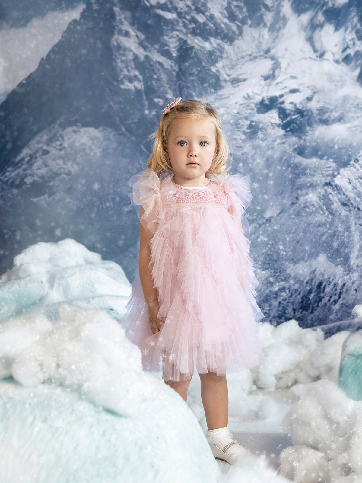 A young girl wears the Bébé Summit Sparkle Tulle Dress by Tutu du Monde, standing amid artificial snow with a snowy mountain backdrop.