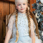 Young girl with long blonde braids wears the Tutu du Monde Diamond Drift Cafe dress and a pearl headpiece, sitting near a Christmas tree by a wooden wall.