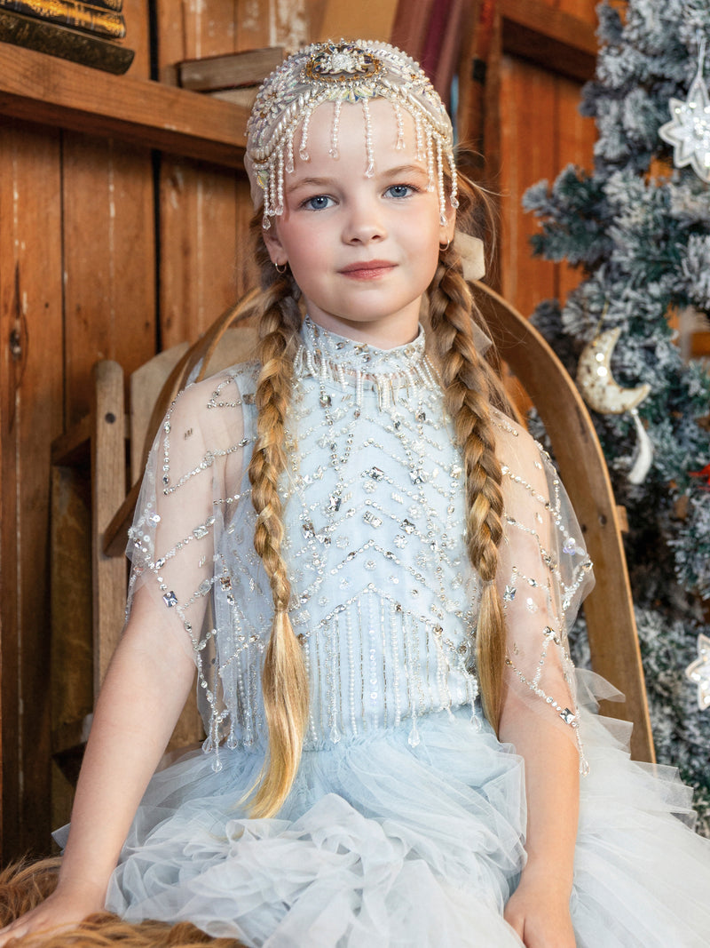 Young girl with long blonde braids wears the Tutu du Monde Diamond Drift Cafe dress and a pearl headpiece, sitting near a Christmas tree by a wooden wall.