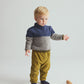 A young child with blond hair stands on a white floor, wearing the Cozmo Baby Boys’ Tigris Knit Sweater in blue and gray, mustard yellow pants, and dark socks. A small wooden toy lies on the floor nearby.