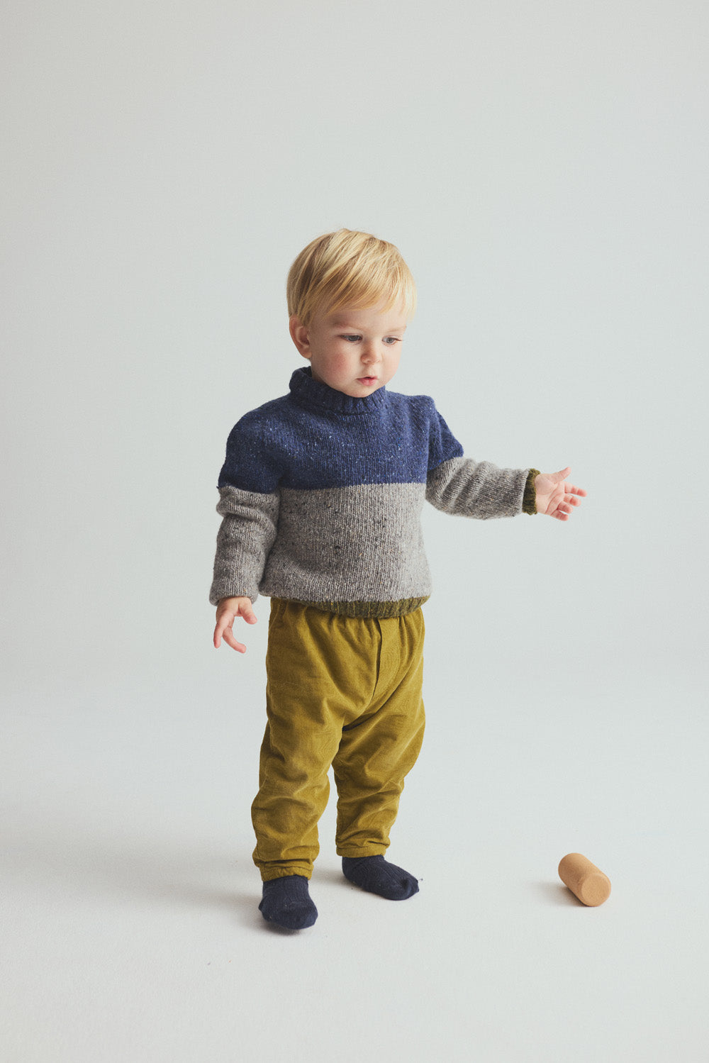 A young child with blond hair stands on a white floor, wearing the Cozmo Baby Boys’ Tigris Knit Sweater in blue and gray, mustard yellow pants, and dark socks. A small wooden toy lies on the floor nearby.