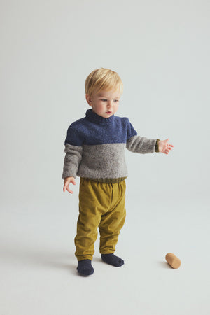 A young child with blond hair stands on a white floor, wearing the Cozmo Baby Boys’ Tigris Knit Sweater in blue and gray, mustard yellow pants, and dark socks. A small wooden toy lies on the floor nearby.