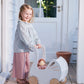 A young child stands barefoot on a wooden porch, smiling as they push the Tender Leaf Toys Sweet Swan Pram stroller with a doll inside.