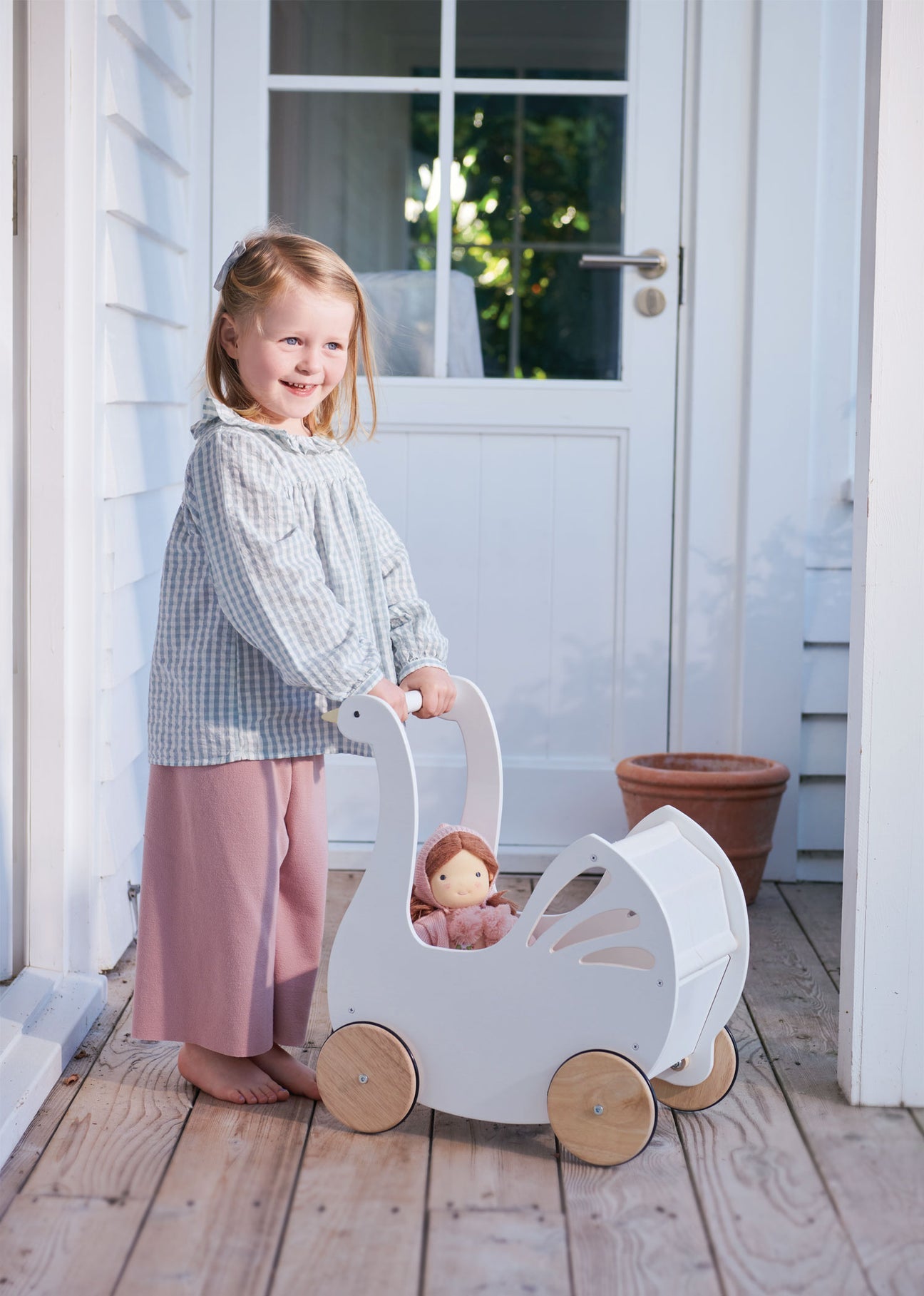 A young child stands barefoot on a wooden porch, smiling as they push the Tender Leaf Toys Sweet Swan Pram stroller with a doll inside.