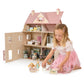 A young girl in a pink skirt plays with wooden dolls and furniture in front of the large, open Alouette House by Tender Leaf Toys, featuring pastel-colored plywood rooms.
