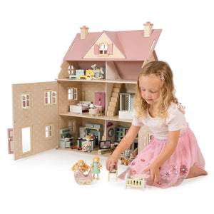 A young girl in a pink skirt plays with wooden dolls and furniture in front of the large, open Alouette House by Tender Leaf Toys, featuring pastel-colored plywood rooms.