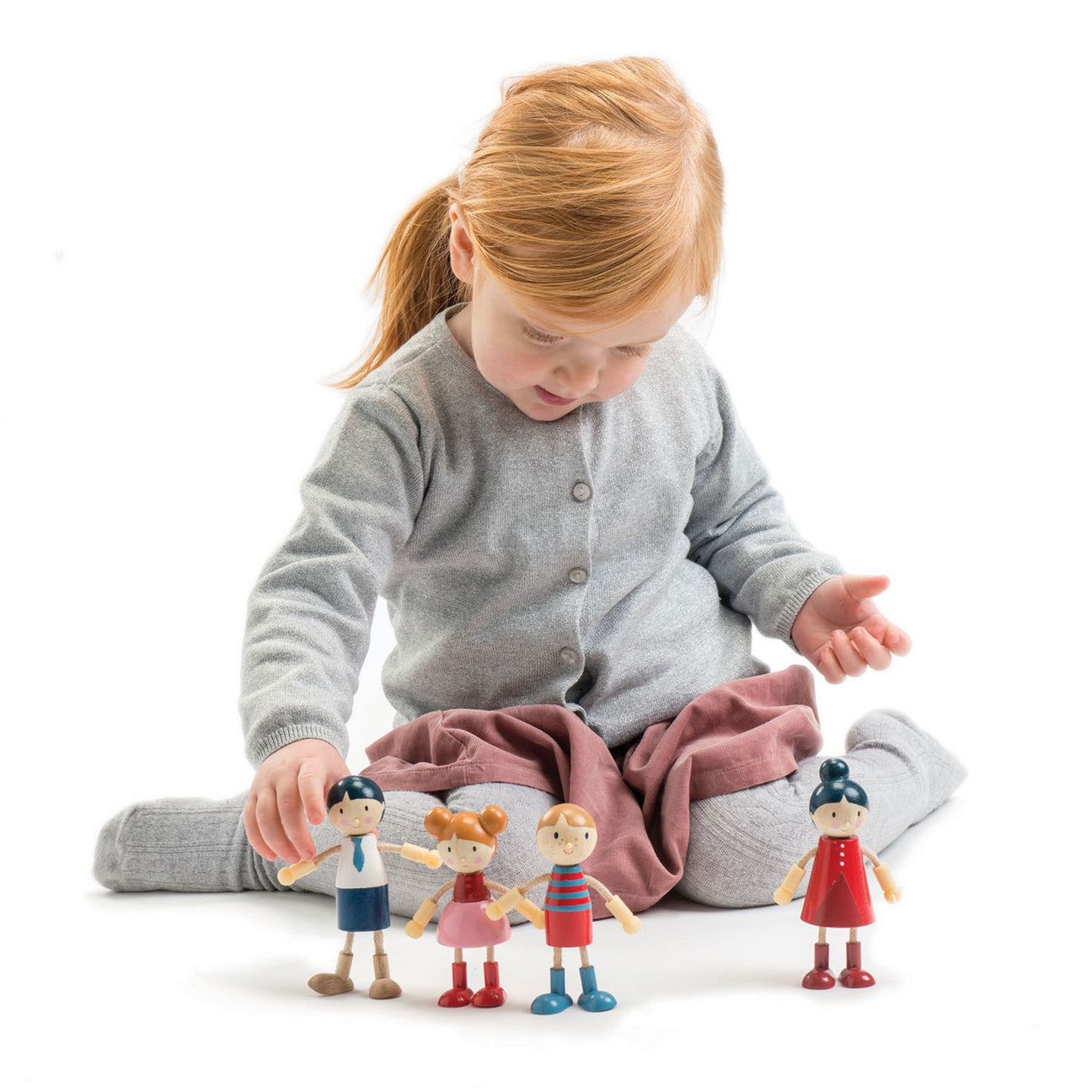 A young child sits cross-legged on the floor, arranging four Tender Leaf Toys Doll Family wooden dolls in front of them, set against a plain white background.